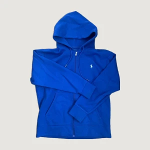 Blå hoodie från Polo Ralph Lauren - Snygg blå hoodie från Polo Ralph Lauren med dragkedja framtill och klassisk huva. Tröjan har långa ärmar, fickor på sidorna och den ikoniska vita logon broderad på bröstet. Perfekt för en chill och sportig look.