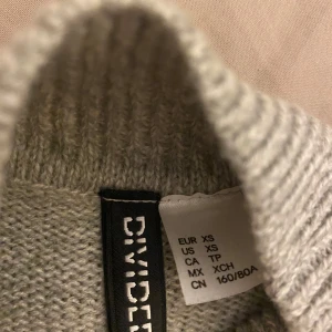 Grå stickad tröja från Divided XS - Mysig grå stickad tröja från Divided i storlek XS. Tröjan har en rak, lite boxig passform med ribbade muddar och högre halsringning. Perfekt för lager-på-lager och chill dagar. Enkel att matcha med jeans eller kjol.