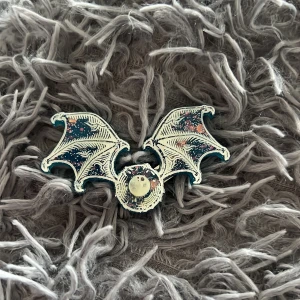 Hårspänne med fladdermusvingar - Unikt hårspänne i form av fladdermusvingar med coolt galaxmönster i blått, vitt och inslag av rosa. Metallklämma på baksidan. Perfekt accessoar för dig som gillar fantasy eller vill sticka ut med din stil.