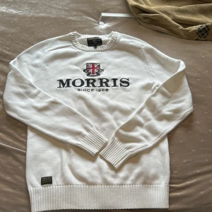 Vit stickad tröja från Morris Gents - Vit stickad tröja från Morris Gents med broderad logga och brittisk flagga på bröstet. Tröjan har rund hals, ribbade muddar och nederkant. Snygg och klassisk stil som passar perfekt till jeans eller chinos.