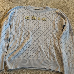 Ljusblå stickad tröja med pärltext - Supersöt ljusblå stickad tröja från Fullamoda med diamantmönster och texten 'chic' i pärlor och gulddetaljer framtill. Tröjan har rund halsringning och långa ärmar, perfekt för dig som gillar unika detaljer och en trendig look.