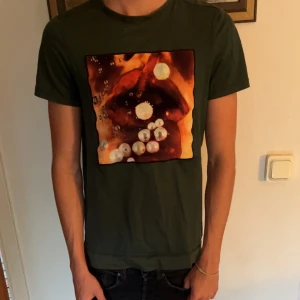 Limitato t-shirt  - Snygg mörkgrön t-shirt med ett stort grafiskt tryck på bröstet i orange, rött och vitt. T-shirten har korta ärmar och en klassisk rund halsringning. Perfekt för dig som vill sticka ut med en cool och unik look.