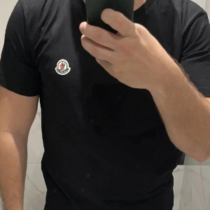 Svart Moncler t-shirt med logga - Svart t-shirt från Moncler med klassisk rund hals och liten broderad logga på bröstet. Enkel och stilren design som passar perfekt till jeans eller shorts. Tillverkad i mjuk bomull för en skön känsla hela dagen.