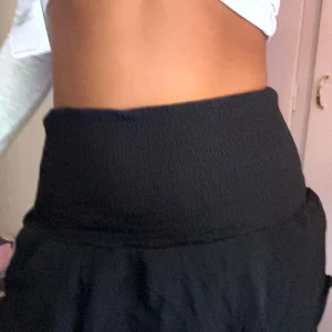 Svart volangkjol - Supersöt svart kortkjol med volanger och hög smockad midja. Kjolen har en flowy känsla och passar perfekt till crop tops eller stickade tröjor. Materialet känns lätt och luftigt, vilket gör den extra skön att bära under varmare dagar.