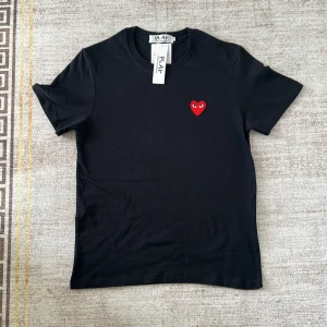 Svart Comme des Garçons Play t-shirt - Svart t-shirt från Comme des Garçons Play med klassisk rund hals och korta ärmar. På bröstet sitter det ikoniska röda hjärtat med ögon, broderat som en snygg detalj. T-shirten är i mjuk bomull och har en clean, stilren look.
