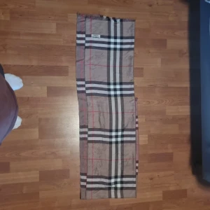 Rutig halsduk i kashmir från Burberry - Snygg rutig halsduk från Burberry i 100% kashmir. Klassisk beige bas med svarta, vita och röda ränder. Mjuk och lyxig känsla, perfekt för att lyfta din outfit med en ikonisk accessoar. Tillverkad i Skottland.