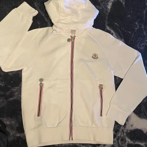 Vit hoodiejacka från Moncler - Anledning för att jag säljer den är för att ja fick den nyligen som en sen födelsedags present och kan nu inte lämna tillbaks tröjan, så måste sälja den en till anledning är att den är för liten på mig. BESKRIVNING:                                Vit hoodiejacka från Moncler med dragkedja och klassiska Moncler-detaljer. Jackan har röda, vita och blå ränder längs dragkedjorna och broderad logga på bröstet. Tillverkad i mjuk bomull och har två fickor framtill. Perfekt för en clean och sportig look