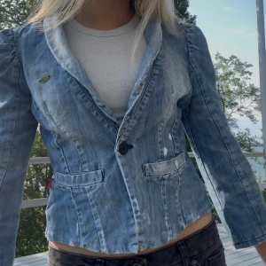 Jeanskavaj med puff-axlar - Säljer en snygg ljusblå jeanskavaj med markerade axlar och figurnära passform. Kavajen har klassisk krage, en knapp framtill och två fickor med lock. Perfekt för dig som vill ha en cool twist på den klassiska kavajen.