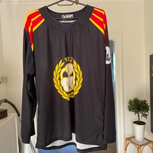 Brynäs tröja - Säljer en svart långärmad Brynäs tröja från säsong 21/22. Tröjan har röda och gula detaljer på axlarna och ett stort klubbmärke i gult och vitt på bröstet. Tillverkad i lätt och ventilerande polyester, perfekt för match eller träning.