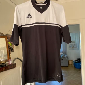 Träningströja - Snygg träningströja från Adidas i svart och vitt med klassisk V-ringning och korta ärmar. Tillverkad i lätt och ventilerande Climalite-material som håller dig torr. Adidas-logga på bröstet och stilren design med vit panel upptill.