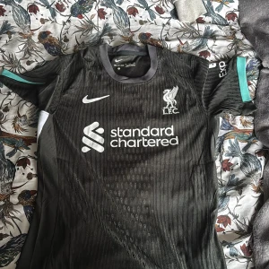 Liverpool svart bortatröja Nike - Snygg svart Liverpool FC bortatröja från Nike med vita loggor och sponsortryck. Tröjan har korta ärmar med turkosa detaljer och är tillverkad i lätt, ventilerande funktionsmaterial. Perfekt för dig som vill visa ditt stöd för LFC på match eller träning.