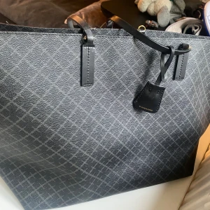 Svart shoppingväska By Malene Birger - Stilren svart shoppingväska från By Malene Birger med rutigt mönster och snygga svarta handtag. Väskan är rymlig och har en klassisk form, perfekt för att bära med sig allt man behöver. Tillverkad i tåligt syntetmaterial med detaljer i svart. Större modellen.  Handtagen är limmade och funkar ok men borde kanske bytas ut till framtiden.