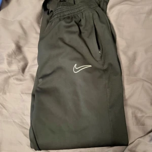 Svarta Nike academy byxor dri-fit - Svarta mjukisbyxor från Nike i storlek XL med Dri-FIT-material som andas. Byxorna har elastisk midja, dragkedjeficka och klassisk vit Nike-logga på benet. Perfekta för träning eller chill, med smalare passform och sportig look. Har blivit för små, köpta från nike.com