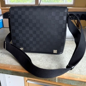 Louis Vuitton svart axelväska Damier - Snygg svart axelväska från Louis Vuitton med klassiskt Damier-rutmönster. Väskan har justerbar axelrem i textil och detaljer i skinn. Perfekt storlek för vardagens essentials och stilren look med silverfärgade metalldetaljer.