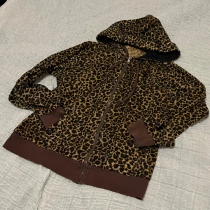 Leopardmönstrad hoodie med huva - Leopardmönstrad hoodie med dragkedja och huva. Tröjan har bruna muddar vid ärmslut och nederkant samt snörning i huvan. Mjuk och skön, perfekt för dig som vill sticka ut lite extra. Långärmad och med praktiska fickor framtill.