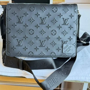 Svart axelväska Louis Vuitton monogram - Säljer en svart axelväska från Louis Vuitton med klassiskt monogrammönster i relief. Väskan har justerbar axelrem med logga, dragkedja på baksidan och detaljer i silver. Tillverkad i skinn med stilren, fyrkantig form och plats för det viktigaste.