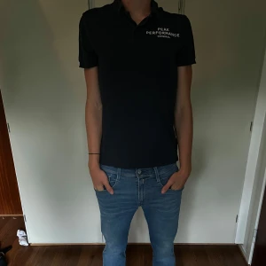 Svart Peak Performance t-shirt - Svart t-shirt från Peak Performance med vit logga på bröstet. Klassisk passform med korta ärmar och krage. Perfekt för en clean och sportig stil. Tillverkad i mjukt material som känns skönt mot huden.