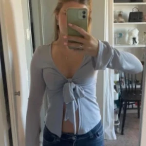 Ljusgrå långärmad topp med knyt - Supersnygg ljusgrå långärmad topp med djup v-ringning och knyt framtill. Modellen är croppad och har en mjuk, stretchig känsla. Perfekt att styla med jeans eller kjol för en trendig look.