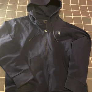 Marinblå/navy hoodie från Polo Ralph Lauren - Snygg marinblå/navy hoodie från Polo Ralph Lauren med dragkedja och klassisk vit broderad logga på bröstet. Tröjan har huva med snörning, två fickor fram och är tillverkad i mjuk bomull. Perfekt för en chill och stilren look.