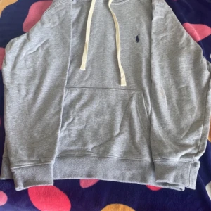 Grå hoodie från Polo Ralph Lauren - Snygg grå hoodie från Polo Ralph Lauren med klassisk broderad logga på bröstet. Tröjan har huva med vita snören, känguruficka och ribbade muddar. Perfekt för chill dagar och enkel att matcha med jeans eller joggers.