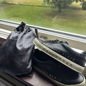 Philipp Plein svarta sneakers med nitar - Svarta sneakers från Philipp Plein i skinn med vita sulor och coola silverfärgade nitar längs sidorna. Skorna har snörning och tydlig logga på sidan samt metallplatta på plösen. Grymma detaljer för dig som vill sticka ut.