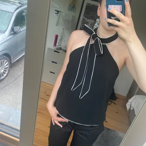Svart halterneck topp med rosett från H&M - Supersnygg svart halterneck topp från H&M med vit kant och stor knytrosett i halsen. Toppen är ärmlös och har öppen rygg, vilket ger en stilren och trendig look. Endast använd en gång av mig❤️