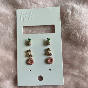 Söta örhängen med frukt och donut - Tre par örhängen från okänt märke och metall, ananas med grönt och guld, körsbär med rosa stenar och guld, samt rosa donut med strössel. Perfekta för dig som gillar lekfulla och färgglada accessoarer. Alla är små och i metall med emalj och stenar. (Ej från H&M, bara hängt dom på pappret). 