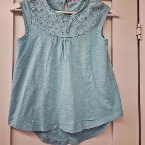 Ljusblå ärmlös topp från Polarn O. Pyret - Supersöt ljusblå ärmlös topp från Polarn O. Pyret med broderade blommor upptill och rund halsringning. Toppen har en lätt A-linjeform och knapp i nacken. Perfekt för varma dagar och enkel att matcha med jeans eller shorts.