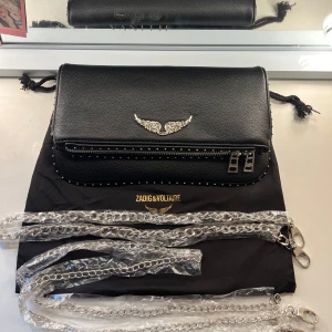 Svart Zadig&Voltaire väska - Snygg svart axelväska från Zadig & Voltaire i skinn med silverfärgade detaljer, kedjerem och coolt vingemblem framtill. Väskan har dragkedja med unika metalldetaljer och små nitar längs kanterna. Perfekt för dig som gillar edgy och stilren design.
