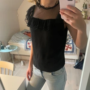 Svart blus med volang och mesh - Supersnygg svart blus med transparent mesh upptill och små prickar. Volangdetaljer vid axlar och halsringning ger en cool look. Ärmlös modell med knäppning bak i nacken. Perfekt för dig som gillar unika detaljer och stilren design.