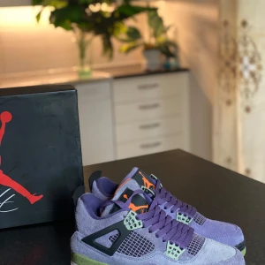 Nike Air Jordan 4 Retro Purple - Unika Nike Air Jordan 4 Retro sneakers i lila mocka med gröna och svarta detaljer samt orange insida. Skorna har klassisk snörning, meshpaneler och robust sula. Perfekta för dig som vill sticka ut med färg och stil. Ikonisk Jumpman-logga på hälen och tungan.