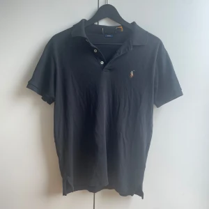Ralph Lauren Piké - Klassisk svart pikétröja från Ralph Lauren i slim fit. Tröjan har korta ärmar, krage och knäppning med tre knappar framtill. Ikoniska broderade polospelaren på bröstet. Tillverkad i 100% bomull för en skön och luftig känsla. Fint skick utöver lappen som är borta. Nypris 1800 kr. 
