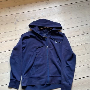 Marinblå hoodie från Polo Ralph Lauren - Säljer en marinblå hoodie från Polo Ralph Lauren i storlek S. Tröjan har dragkedja, huva med snörning och klassisk logga på bröstet. Tillverkad i mjuk bomull och har praktiska fickor framtill. Perfekt för en chill och stilren look. Har en defekt som visas i tredje bilden därav priset
