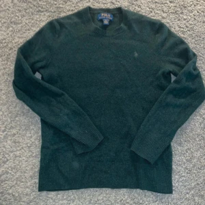 Mörkgrön stickad tröja Polo Ralph Lauren - Mörkgrön stickad tröja från Polo Ralph Lauren med klassisk rund hals och broderad logga på bröstet. Tröjan har ribbade muddar vid ärmslut och nederkant, perfekt för lager-på-lager under hösten och vintern.