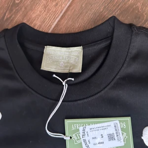 Svart blommig t-shirt från Denim Tears - Svart t-shirt från Denim Tears med stora vita blommiga tryck över hela plagget. Klassisk rund hals och korta ärmar. Tillverkad i mjuk bomull som känns skön mot huden. Perfekt för dig som vill sticka ut med en cool och unik stil. Kläd plagget kommer med pris lappen 