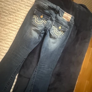 True Religion Joey Low Rise Flare Jeans - Säljer ett par  low waist blå True Religion Joey jeans med vit detalj på fickorna. I ett bra skick, men då de inte passar mig längre så säljer jag dem. Kanske några få tecken på användning men inget som märks av💕 i storlek 24, fråga för mått!