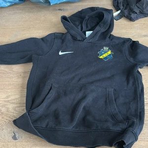 Svart AIK hoodie från Nike - Svart hoodie från Nike med AIK-logga på bröstet och klassisk känguruficka. Tröjan har huva och är tillverkad i mjuk bomullsmix, perfekt för dig som vill visa ditt AIK-stöd. Snygg och sportig stil med långa ärmar och bekväm passform.