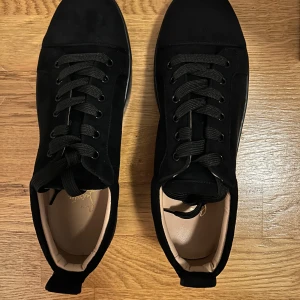 Svarta sneakers från Christian Louboutin - Snygga svarta sneakers från Christian Louboutin med klassisk röd sula. Skorna har snörning och är tillverkade i mocka. Perfekta för en stilren look. Ingår kartong och autentisering. Använda i nyskick och är storlek 41