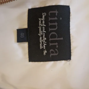 Vit quiltad pufferjacka från Tindra - Snygg vit pufferjacka från Tindra i storlek 40. Jackan har quiltad design, guldiga dragkedjor och en mysig huva med mjukt foder. Perfekt för kyliga dagar när du vill hålla stilen och värmen. Långärmad och med praktiska fickor framtill.