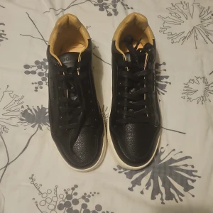 Svarta sneakers från Björn Borg - Snygga svarta sneakers från Björn Borg med beige detaljer vid hälen och insidan. Skorna har en vit platt sula, snörning och är tillverkade i skinn med en lätt texturerad yta. Perfekta för dig som gillar stilrena och sportiga vibes. Ingen låda ingår