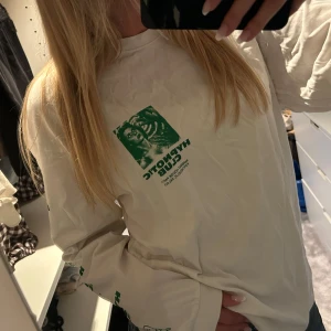 Vit långärmad tröja Hypnotic Club - Vit långärmad tröja från Hypnotic Club med stort grönt tryck framtill och på ärmarna. Trycket har retroinspirerad grafik och text med grodor och klubbtema. Tröjan har en avslappnad passform och är gjord i mjuk bomull. Perfekt för dig som gillar unika statement-plagg.