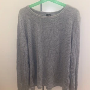 Grå ribbad tröja från Vero Moda - Grå långärmad tröja från Vero Moda i ribbat material med diskret mönster. Tröjan har en rund halsringning och en enkel, clean look som passar perfekt till jeans eller kjol. Skön och lätt att bära under höst och vinter.