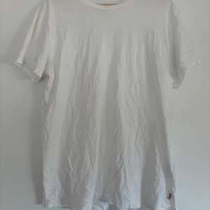 Vit slim t-shirt från Levi's - Vit t-shirt från Levi's i slim fit-modell. Klassisk rund hals och korta ärmar, tillverkad i mjuk bomull. Diskret Levi's-röd etikett på sidan ger en clean och stilren vibe. Perfekt för dig som gillar enkel och snygg stil.