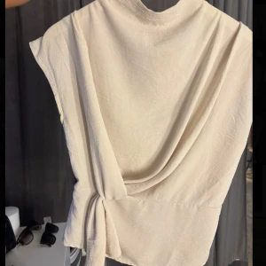 Beige omlottopp från Boohoo - Säljer min beige omlottopp från Boohoo i storlek 36. Toppen har knyt i sidan, ärmlös design och lätt rynkad midja med volangkant💕Endast använd en gång!!