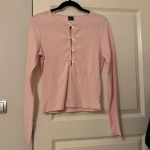 Ljusrosa ribbad topp med rosetter - Oanvänd ljusrosa långärmad topp från Gina Tricot med ribbad struktur och fyra små rosetter framtill. Säljer då den tyvärr är för liten. Storleken är L men skulle säga att den sitter som en M