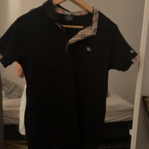 Svart Burberry t-shirt med rutig krage - Svart t-shirt från Burberry London med klassisk rutig krage och detaljer vid ärmslut. Ikonisk Burberry-logga broderad på bröstet. T-shirten har korta ärmar och knappslå framtill. Perfekt för dig som gillar stilrena designer med en twist.