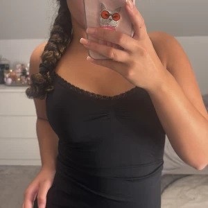 Svart linnetopp med spetskant - Svart linnetopp med smala axelband och söt spetskant längs v-ringningen. Toppen har en liten rynkdetalj (scrunch) framtill och är figurnära i modellen. Perfekt att styla med jeans eller kjol för en enkel men snygg look.