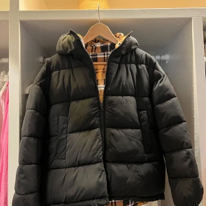 Vändbar pufferjacket från Stay - Vändbar & varm pufferjacket från Stay i storlek XS. Ena sidan är helsvart och andra sidan har ett rutigt mönster i gult, svart och vitt. Jackan har huva, dragkedja, fickor och snörning nertill för att justera passformen. Perfekt för dig som vill variera din stil.