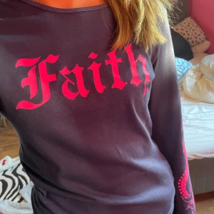 Mörkblå långärmad Faith-tröja - Cool långärmad tröja i mörkblått med starkt rosa tryck. Framsidan har stor text 'Faith' i gotisk stil och baksidan har ett fett motiv med dödskalle och orm. Tryck även på ärmarna. Tröjan är gjord i mjuk bomull och har en normal passform. Perfekt för dig som gillar edgy streetstyle.