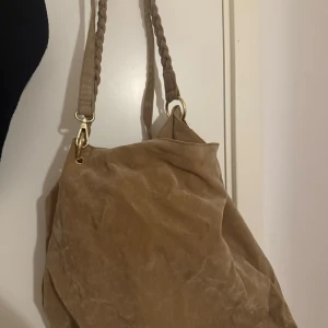 Brun/beige axelväska i mocka från Gina Tricot - Stilren beige axelväska från Gina Tricot i mjuk mocka med flätade axelband och guldfärgade metalldetaljer. Rymlig insida med innerficka och dragkedja. Perfekt för dig som vill ha en snygg och praktisk väska till vardagen.